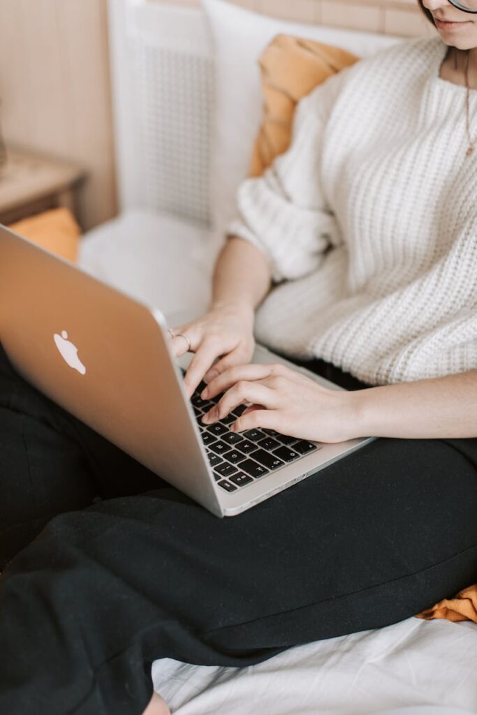 A woman in a sweater typing on a laptop while sitting on a bed in a cozy bedroom environment.